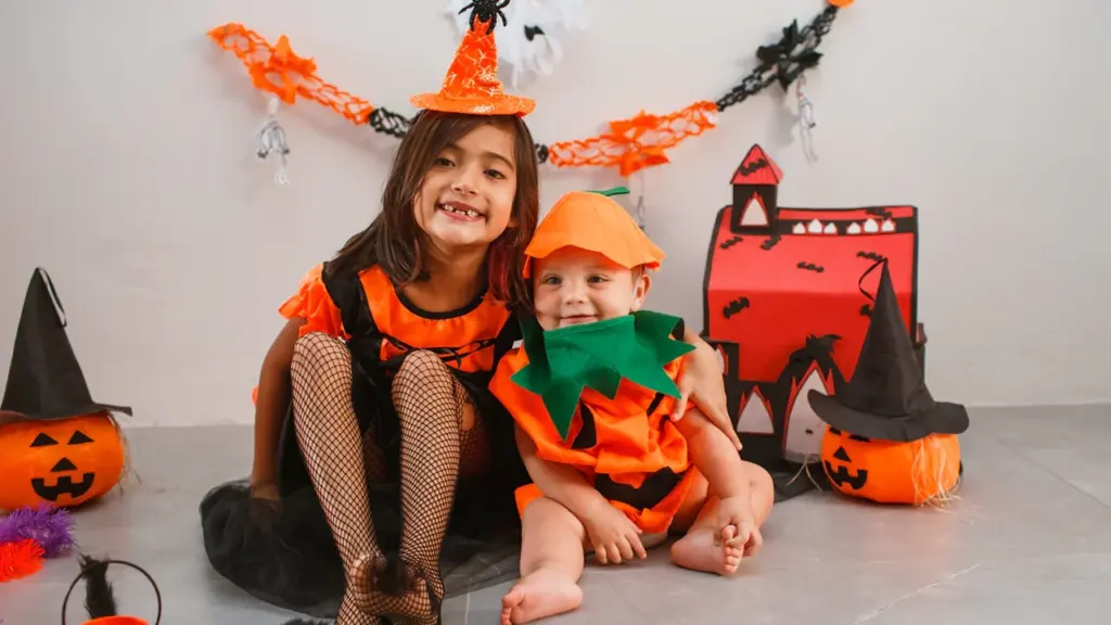 Zwei Kinder in Halloween-Kostümen sitzen fröhlich gemeinsam vor einer herbstlichen Dekoration.